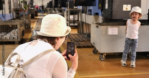 Woman takes photo of boy in front of machine on assembly line. Child turns on the spot and talks. Mother and son visit the car factory.