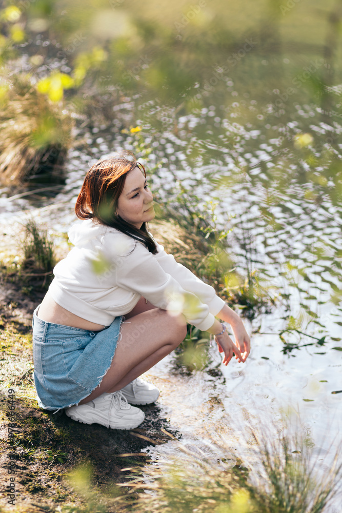 Portrait of a young woman with red hair through the leaves of the trees. She squats near a tranquil lakeside to enjoing the nature.
