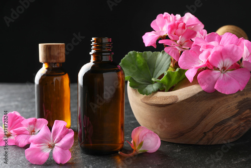 Bottles of geranium essential oil, beautiful flowers and mortar with pestle on black table, closeup