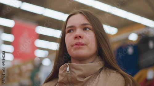 Close-up of a young girl in a brown jacket, looking around in a mall. The background is blurred, emphasizing her curious and observant expression