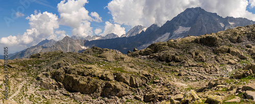 A panoramic view of a rugged mountain landscape featuring rocky terrain and patches of greenery under a clear blue sky with scattered clouds