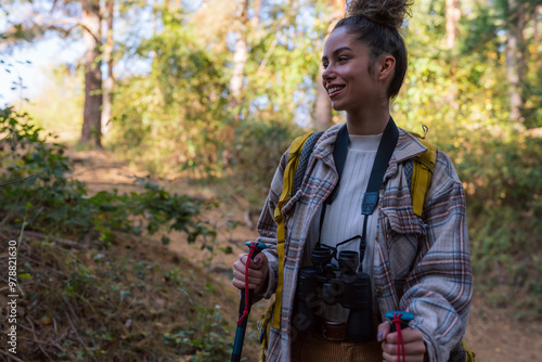 A young woman with curly hair in her 20s exploring the forest trail during autumn, wearing warm clothing and equipped with a yellow backpack, trekking poles, and binoculars.