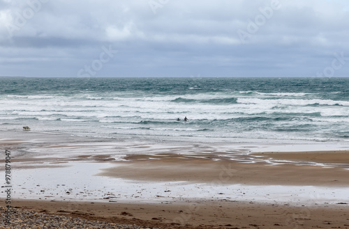 Waves crash on a sandy beach in Strandhill, Ireland, under a cloudy sky with two surfers in the water and a dog running along the shore