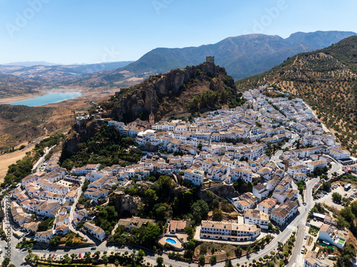 Aerial view of picturesque whitewashed buildings and historic castle on a hillside overlooking a serene lake, Zahara de la Sierra, Spain.