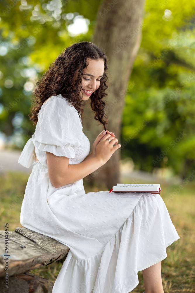 Naklejka premium Laughing woman sitting on a wooden bench in a meadow under a tree in summer. Young girl in white dress relaxing studying and reading a book in nature on vacation
