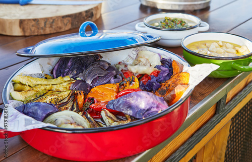 A colorful dish with roasted vegetables including bell peppers, zucchini, red cabbage, and onions placed in a vibrant red pot on a wooden table photo