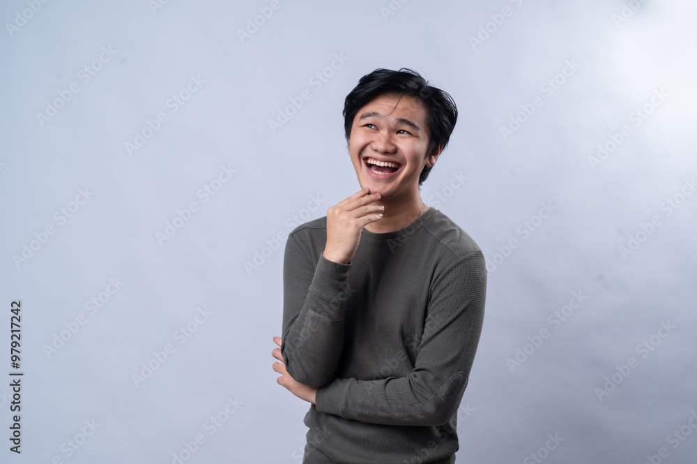 An Asian man wearing a casual long-sleeve gray shirt is standing with a thoughtful expression, hand resting under his chin, as if deep in thought or contemplating an idea. White background An Asian man wearing a casual long-sleeve gray shirt is standing with a thoughtful expression, hand resting under his chin, as if deep in thought or contemplating an idea. White background