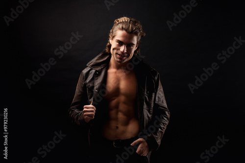 Handsome guy fashion model in unbuttoned leather jacket with abs muscle press posing in black studio shadow, strong look. Perfect young man with lovely muscular sexy torso, image. Copy ad text space