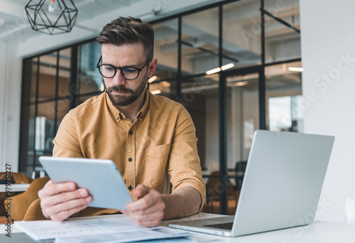Serious businessman inside the office, utilizing a tablet and laptop to analyze data while reviewing online financial reports, showing deep concentration.