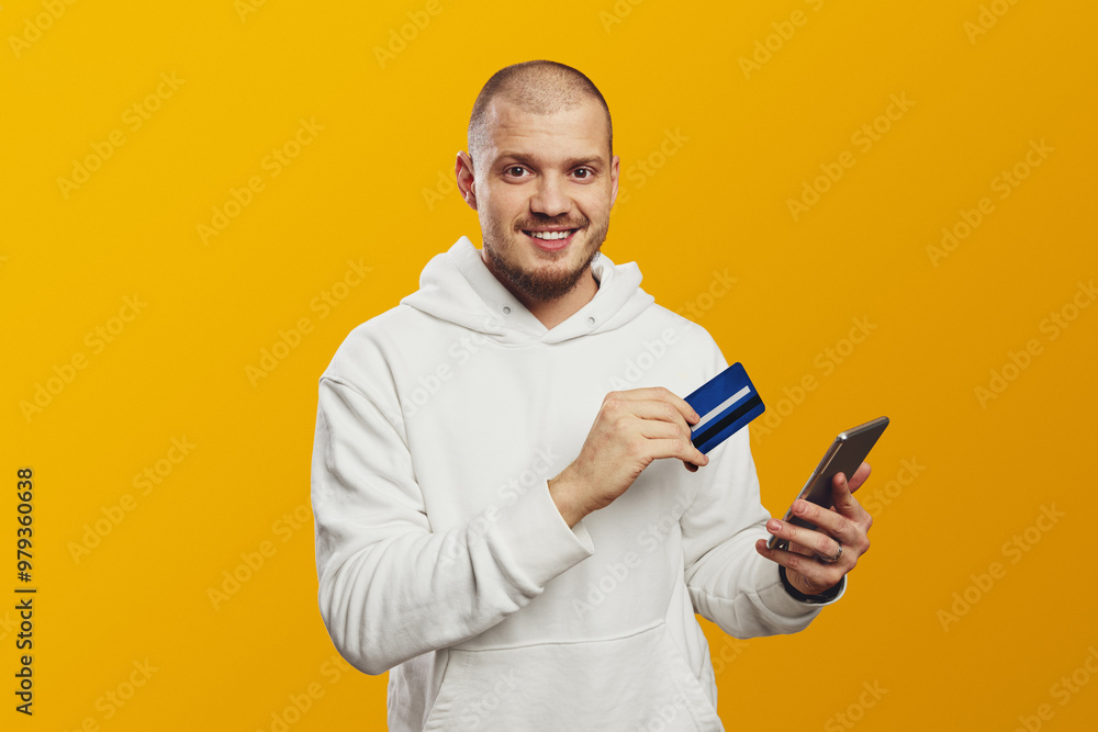 Attractive handsome guy in white hoodie holds credit card and smartphone isolated on yellow background. Online shopping. Online payment. Attractive handsome guy in white hoodie holds credit card and smartphone isolated on yellow background. Online shopping. Online payment.