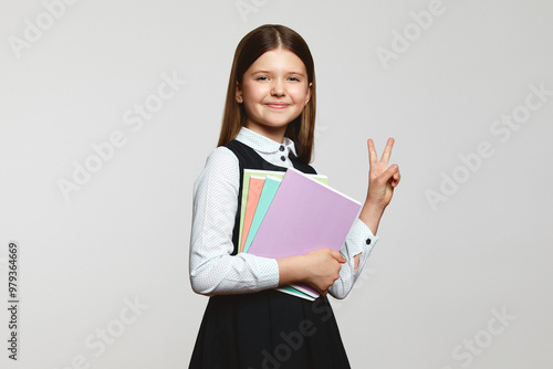 Excited girl in school uniform looking at camera while holding textbooks and showing peace gesture against white background. Back to school concept