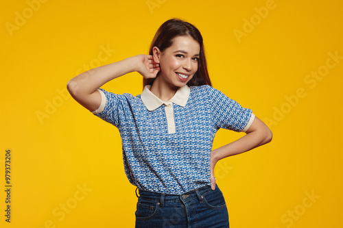 Happy carefree young girl with joyful expression, smiling while standing in relaxed pose against yellow background, holding hand on waist