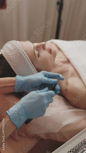 Aesthetician in blue gloves prepares a derma pen for a microneedling procedure on a female client in a spa setting.