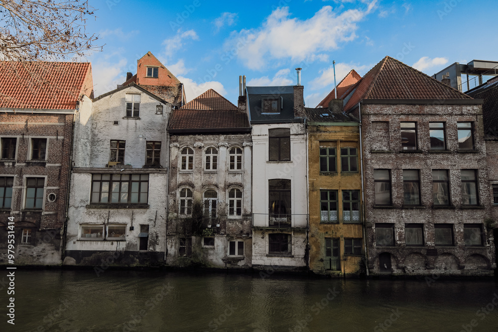 Fototapeta premium Historic buildings along the waterway in Ghent, Belgium, showcasing architectural diversity under a clear blue sky