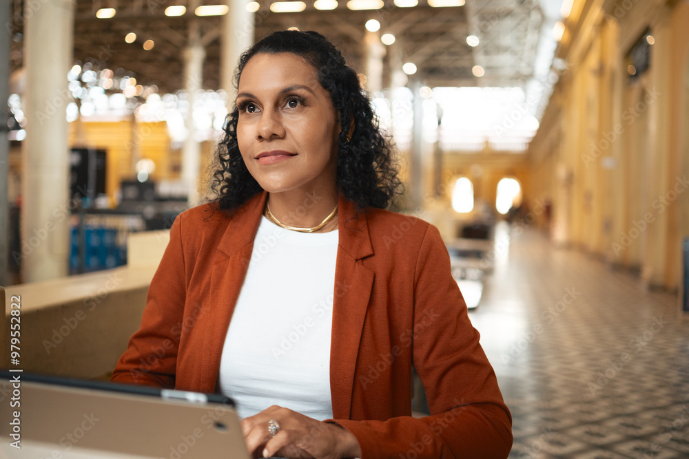 Fototapeta premium Determined Black businesswoman working remotely in modern marketplace. Focused professional in business attire using laptop in bright public space..