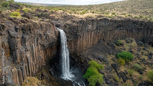 Waterfall tumbling down a volcanic rock face, surrounded by rugged terrain and sparse vegetation.