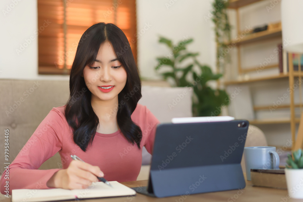 Young businesswoman reading business data on tablet and writing note in paper while working at home Young businesswoman reading business data on tablet and writing note in paper while working at home