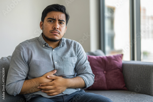 Young man suffering from strong stomach or abdominal pain while sitting on a couch at home photo