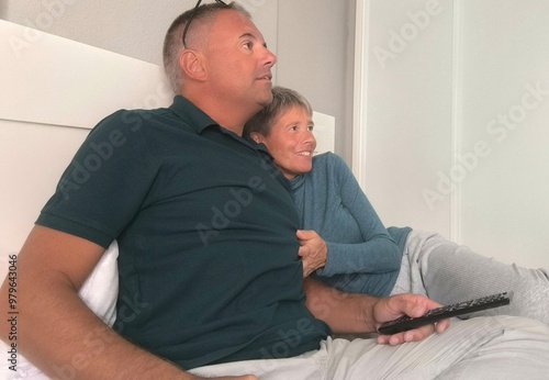 Funny couple watching tv and laughing on a bed in the day.Strange couple watching tv and laughing on the bed. Couple in love, sitting on the bed watching tv. Lovers watching tv chatting in bed.