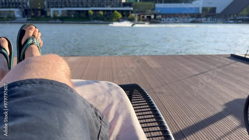 Person relaxing in pool. Man wearing blue shorts and flip-flops is lying and relaxing on a sun lounger on a wooden pier at a tourist resort and looking at jet skis riding on a lake. photo