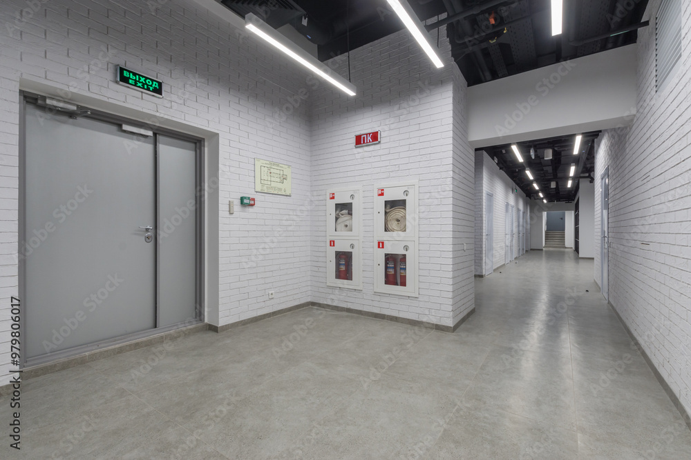 A corridor with white brick walls in an office building. A gray door with a green emergency exit sign. A corridor with white brick walls in an office building. A gray door with a green emergency exit sign.