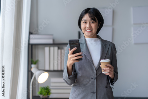 Asian businesswoman is holding a smartphone and a coffee cup while smiling