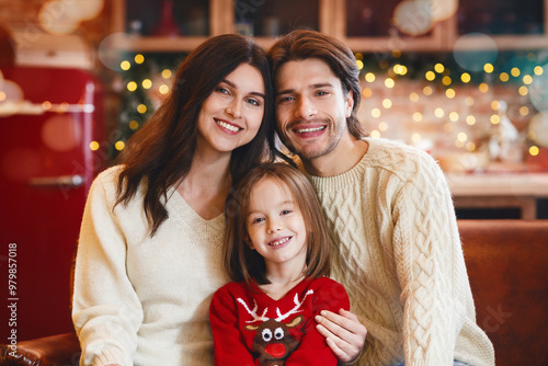 Affectionate little family getting ready for New Year celebration, sitting on sofa, smiling at camera over decorated kitchen, free space