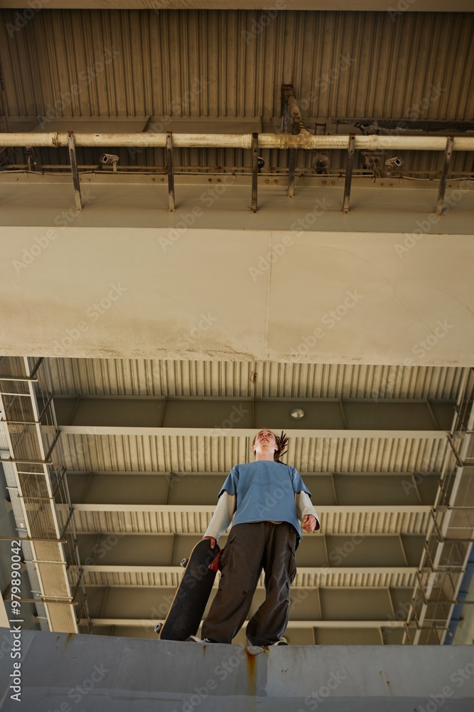 Skateboarder under overpass looking upward while gripping skateboard. Metal structure visible in background, casting shadows onto concrete surface Skateboarder under overpass looking upward while gripping skateboard. Metal structure visible in background, casting shadows onto concrete surface