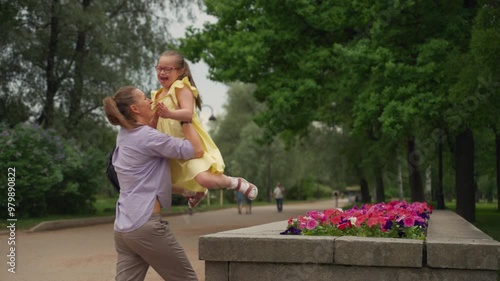 In a summer park, mother helps her young daughter with Down syndrome jump down from a high flowerbed. Both are cheerful and laughing. Scene showcases theme of supporting children with special needs