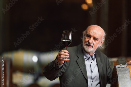 Caucasian senior man in a suit, standing in an old wine cellar with wooden barrels, expertly tasting red wine, checking a color, smell, and taste
