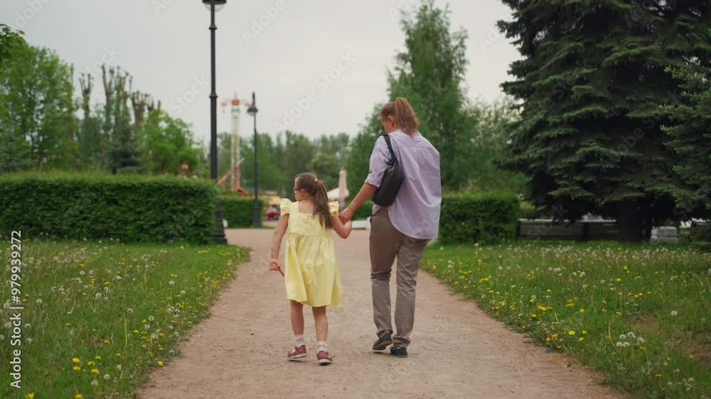Mother and her young daughter with Down syndrome walk hand in hand towards amusement park. Girl is wearing yellow dress, and they are surrounded by green trees and flowers Mother and her young daughter with Down syndrome walk hand in hand towards amusement park. Girl is wearing yellow dress, and they are surrounded by green trees and flowers