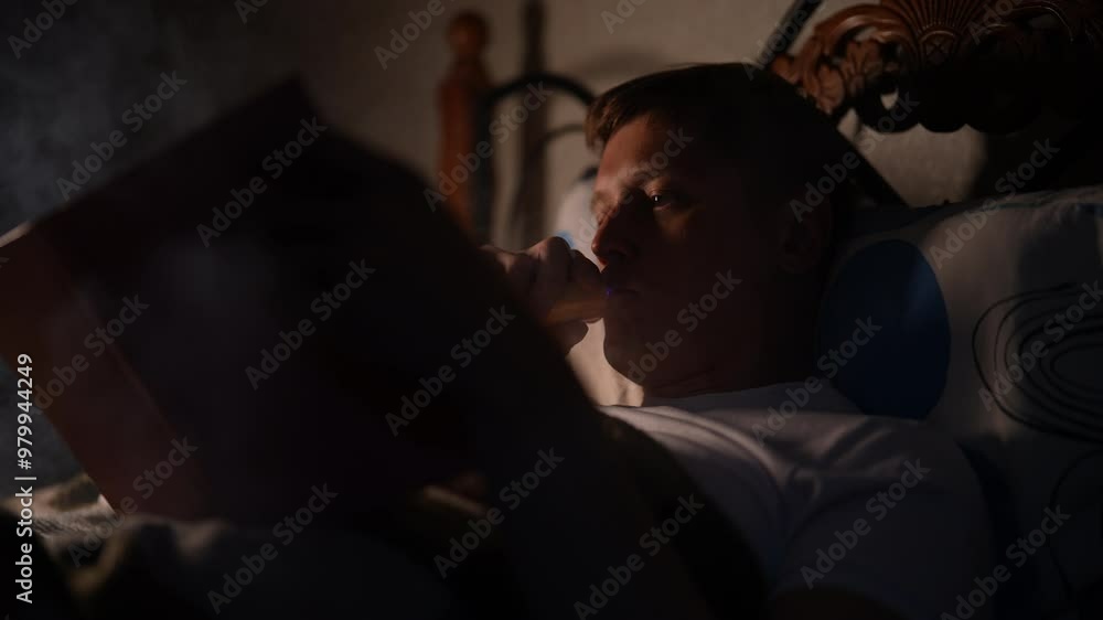 Portrait of young man lying in bed reading book while vaping electronic cigarette late at nigh, at dark bedroom with warm light from bedside lamp illuminating book and creating cozy atmosphere. Portrait of young man lying in bed reading book while vaping electronic cigarette late at nigh, at dark bedroom with warm light from bedside lamp illuminating book and creating cozy atmosphere.