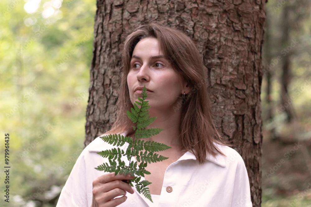 young woman in the forest near a tree holds a fern in her hand young woman in the forest near a tree holds a fern in her hand