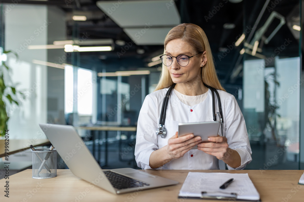 Healthcare professional wearing glasses and stethoscope uses tablet and laptop in modern office for research and communication. Focused medical expert demonstrating technology use in healthcare Healthcare professional wearing glasses and stethoscope uses tablet and laptop in modern office for research and communication. Focused medical expert demonstrating technology use in healthcare