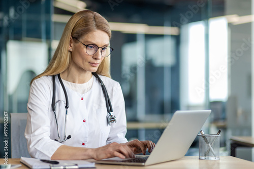 Female medical professional using laptop in office. Doctor wears stethoscope, eyeglasses, focused on work. Depicts healthcare and technology integration. Represents modern medical practice.