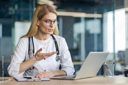 Female doctor wearing stethoscope engaged in online video call using laptop. Medical professional discussing health remotely. Concept of telemedicine, virtual consultation, technology in healthcare.