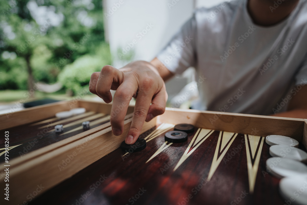 A person playing backgammon outside, focusing on their hand moving a game piece. The setting is a sunny garden, creating a relaxed and fun atmosphere.