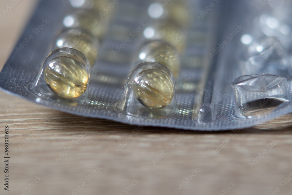 Close-up of soft gel capsules in a blister pack. The capsules appear translucent with a golden hue, set against a wooden background. Close-up of soft gel capsules in a blister pack. The capsules appear translucent with a golden hue, set against a wooden background.