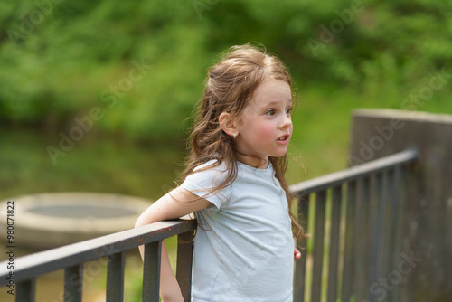 Obraz na płótnie Little preschool girl in a T-shirt leaning on a railing near a pond in a summer park