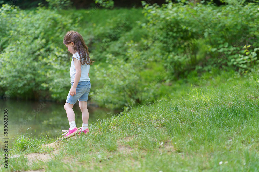 Little cute preschool girl in a T-shirt plays on the background of a pond. Happy childhood concept outdoors Little cute preschool girl in a T-shirt plays on the background of a pond. Happy childhood concept outdoors