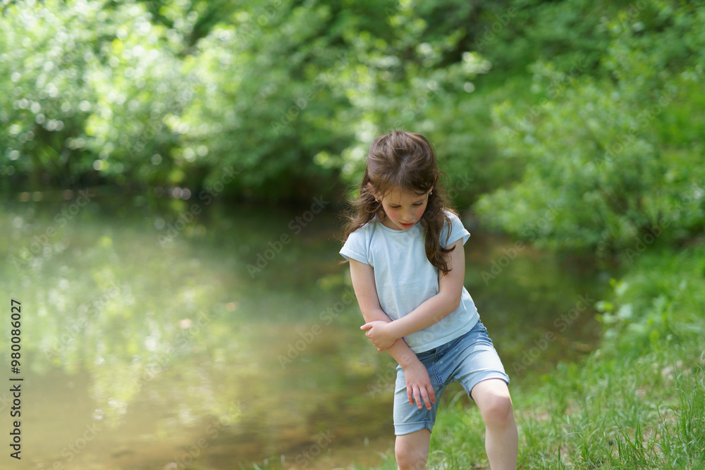 Little cute preschool girl in a T-shirt plays on the background of a pond. Happy childhood concept outdoors Little cute preschool girl in a T-shirt plays on the background of a pond. Happy childhood concept outdoors