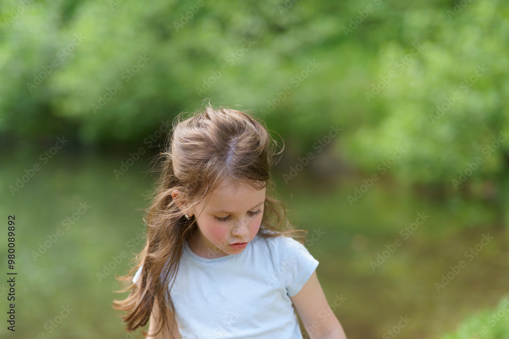 Little cute preschool girl in a T-shirt plays on the background of a pond. Happy childhood concept outdoors Little cute preschool girl in a T-shirt plays on the background of a pond. Happy childhood concept outdoors