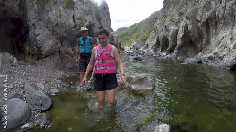 An older man and a young woman wearing life jackets hike through knee deep water in Somoto Canyon An older man and a young woman wearing life jackets hike through knee deep water in Somoto Canyon