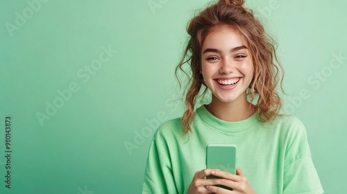 A cheerful young woman holds a smartphone, smiling against a soft green background, embodying joy and modern technology.