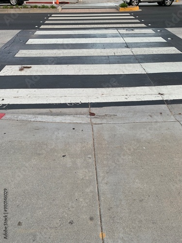 A clear midday view of a classic black-and-white striped crosswalk in Santa Monica, California. The sun casts minimal shadows, highlighting the textures of the worn crosswalk.