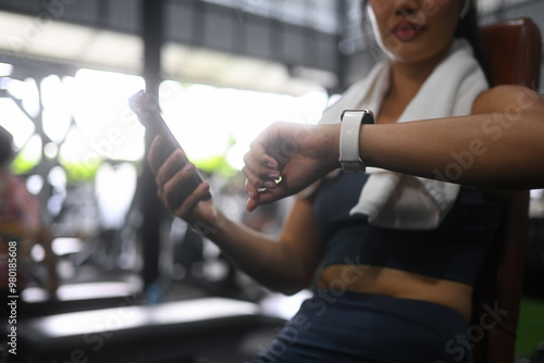 Athletic woman in sportswear sitting on bench in gym and checking fitness tracker