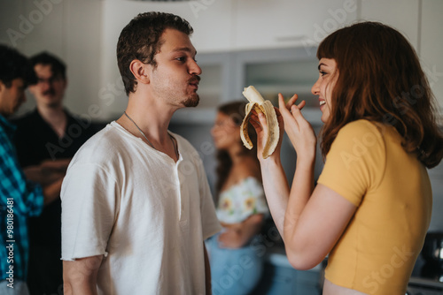 A group of friends share a fun and casual interaction in a modern kitchen. A woman playfully holds a banana while her friend looks amused, capturing a moment of joy and connection.