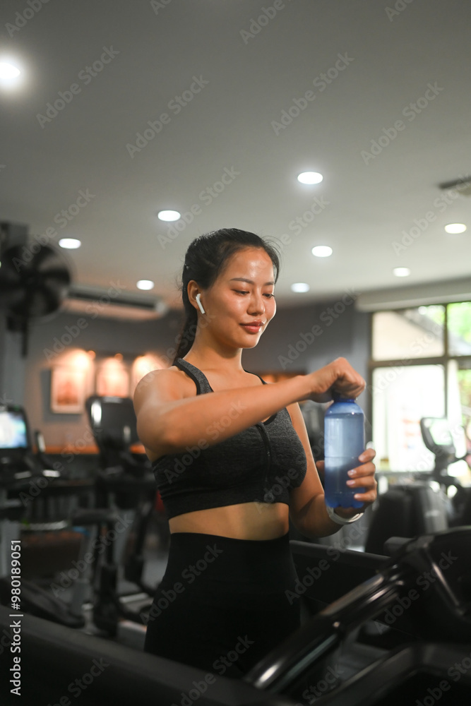Fototapeta premium Smiling fit young woman in sportswear drinking water while walking on a treadmill