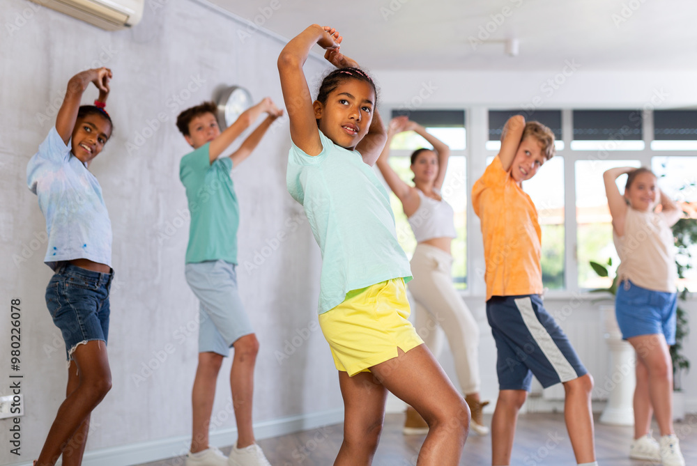 Fototapeta premium Dynamic little girl training Hip hop dance poses in dancehall with other attendees of dancing courses