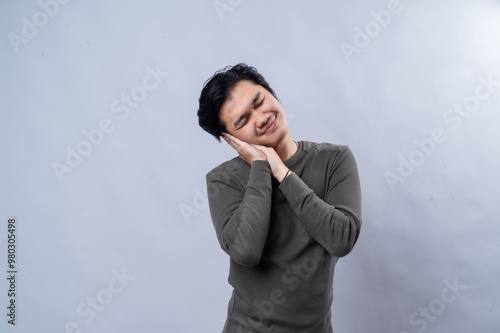 Asian man posing with a soft, pleasant expression, resting his head on his hands as if pretending to sleep or relax. He smiles gently, wearing a dark, long-sleeve shirt. The backdrop is a plain white